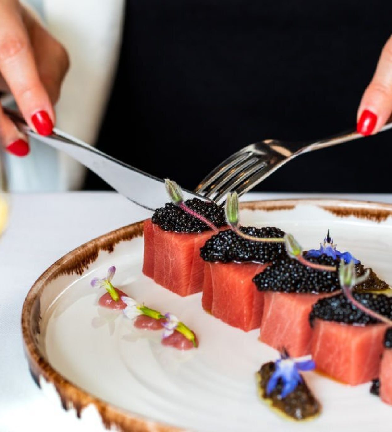 Close up detail of woman having dinner in gourmet restaurant. Macro close up of female hands next to blue fin tuna dish with beluga caviar and a glass of white wine.