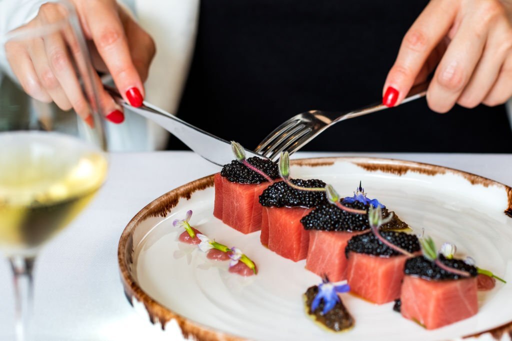 Close up detail of woman having dinner in gourmet restaurant. Macro close up of female hands next to blue fin tuna dish with beluga caviar and a glass of white wine.